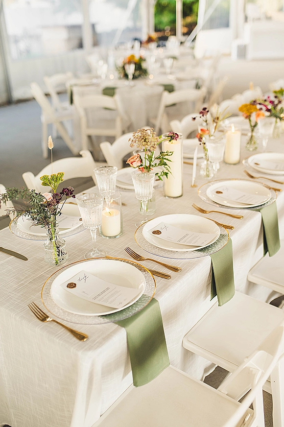 Reception tablescape with wedding place setting, white linens, sage green napkins, gold flatware, wax-seal menus, candles under a tent