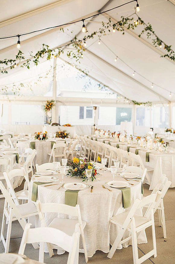 Reception tablescape with round tables, white linens and sage green runners, gold flatware and floral centerpieces under string lights in a tent