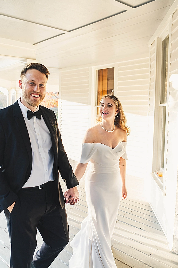 Couple portrait of bride and groom holding hands on a covered porch, her off-shoulder gown with pearl necklace, his black tuxedo and bow tie in daylight