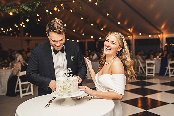 Cake cutting by bride and groom with baby's breath-decorated white cake on a stand, under string lights in a reception tent
