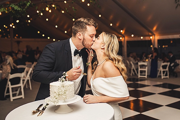 Cake cutting moment as bride and groom kiss beside a buttercream wedding cake with baby’s breath under string lights in a tent reception