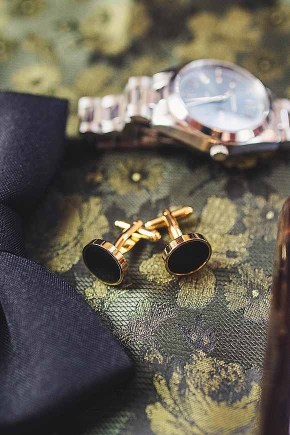 Groom accessories flat lay featuring gold cufflinks and a wristwatch beside a necktie and suit jacket on patterned fabric background