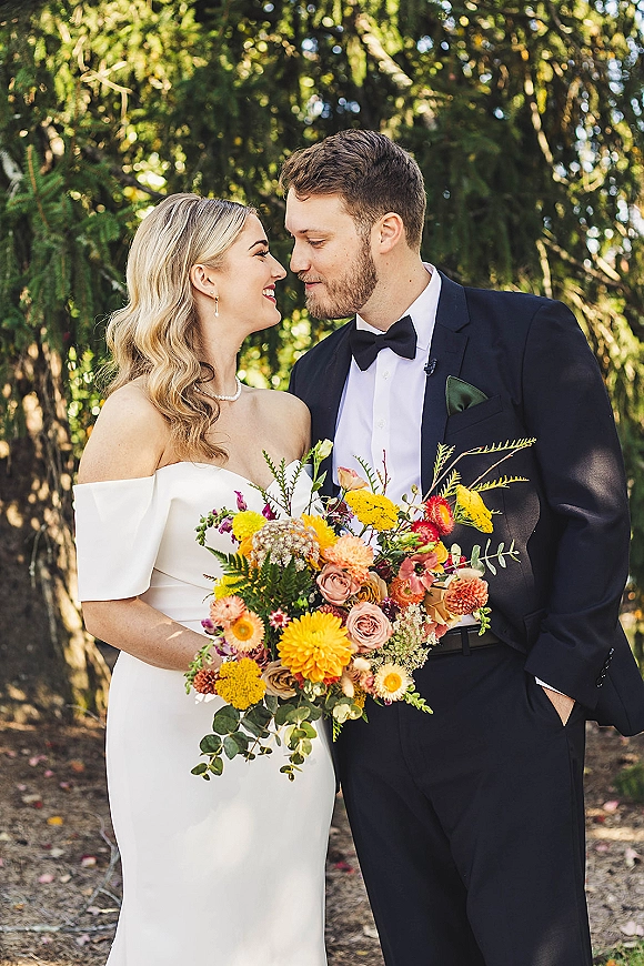 Couple portrait of bride and groom nose to nose, holding a colorful bouquet in dappled sunlight amid evergreen garden trees