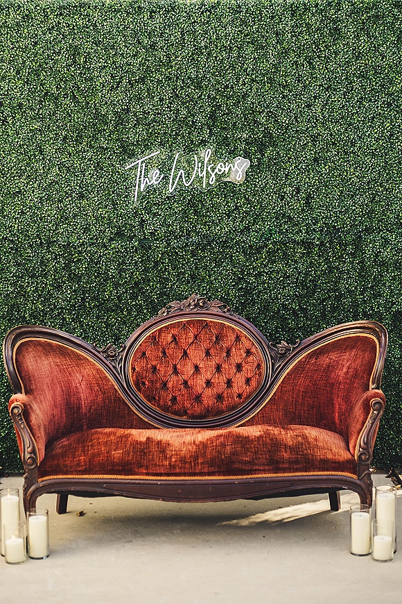 Wedding lounge setup with a vintage velvet sofa, neon name sign on a green hedge wall, and pillar candles on a concrete floor