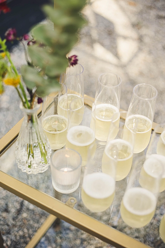 Champagne toast setup with wedding champagne station of coupe glasses on a glass tray table, gold frame, flowers, and a votive candle on a sunlit stone patio