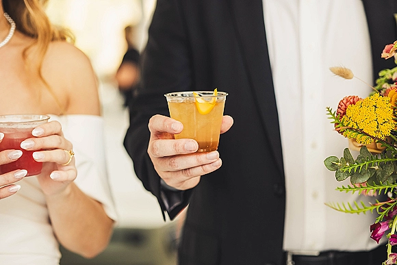 Wedding cocktails held by bride and groom, signature wedding drinks with citrus peel garnish, rings and florals in a blurred reception space