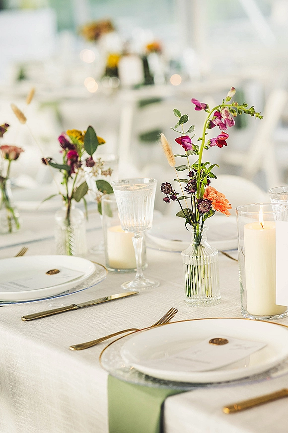 Reception tablescape with wedding table setting, bud vase florals in clear vases and pillar candles on white linens with gold flatware in soft daylight interior
