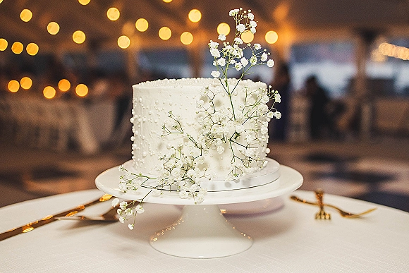 Wedding cake with pearl icing and baby’s breath on a white stand, set on a rustic reception table with string lights bokeh