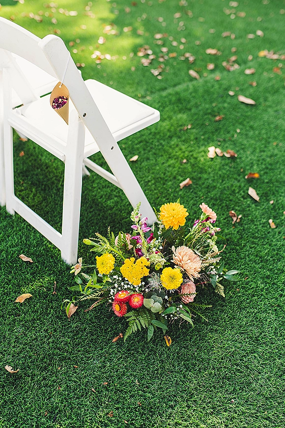 Wedding aisle decor with white folding chairs and chair petal cones, flower petals and greenery on a grassy lawn with scattered leaves