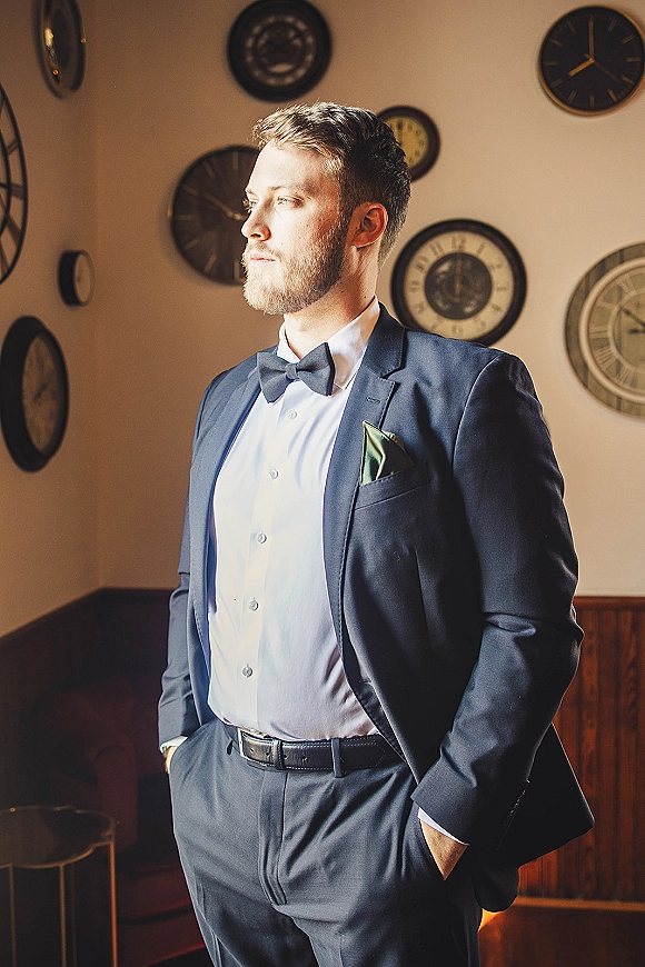 Groom portrait in a navy suit with bow tie and pocket square, hands in pockets by wall clocks, leather sofa, and wood paneling indoors