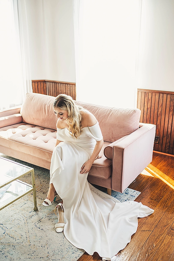 Bridal portrait of a modern bride in an off the shoulder wedding dress sitting on a pink velvet sofa by a sunlit window