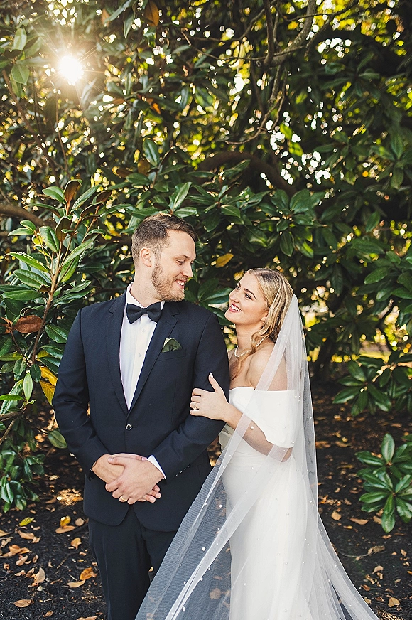 Couple portrait of bride and groom walking a garden path, bride holding his arm in veil and pearls, groom in tuxedo under sun flare