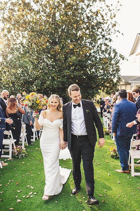 Wedding recessional as bride and groom walk the aisle holding hands, her bouquet raised, guests tossing confetti on a sunny lawn