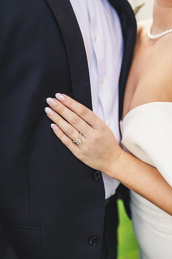 Wedding ring close-up of a diamond engagement ring and band on a nude-manicured hand resting on a groom’s black tuxedo, greenery blurred behind