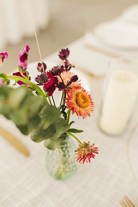 Wedding centerpiece with bud vase centerpiece of pink and orange blooms and eucalyptus, beside a taper candle on a white tablecloth place setting