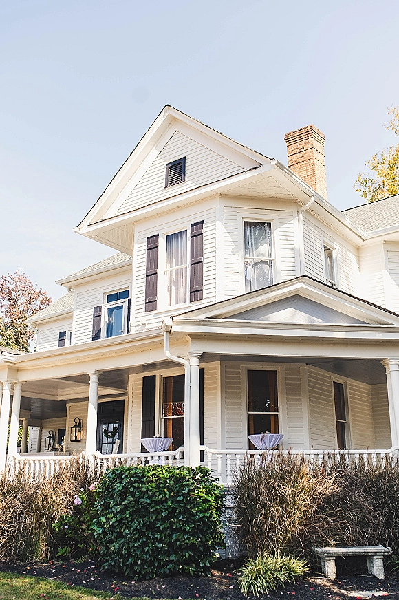 Wedding venue exterior with wraparound porch, black shutters, and high-top cocktail tables with white linens under porch lanterns and blue sky