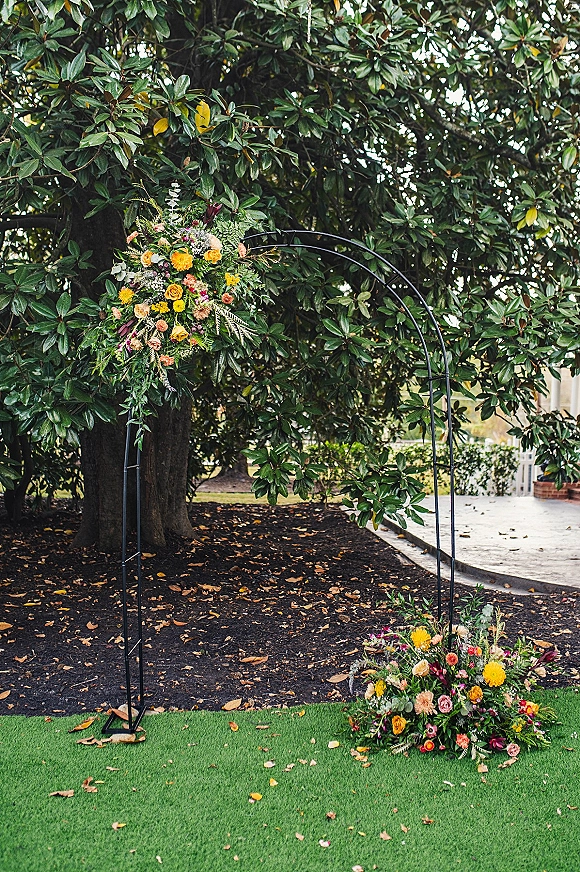 Wedding ceremony arch with an asymmetrical floral arch of colorful blooms and greenery, set on a garden lawn beneath a large tree