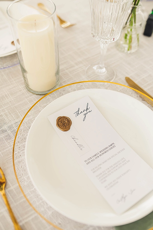 Wedding place setting with gold rim plates, menu card and wax seal stationery, crystal goblet and gold flatware on lace runner