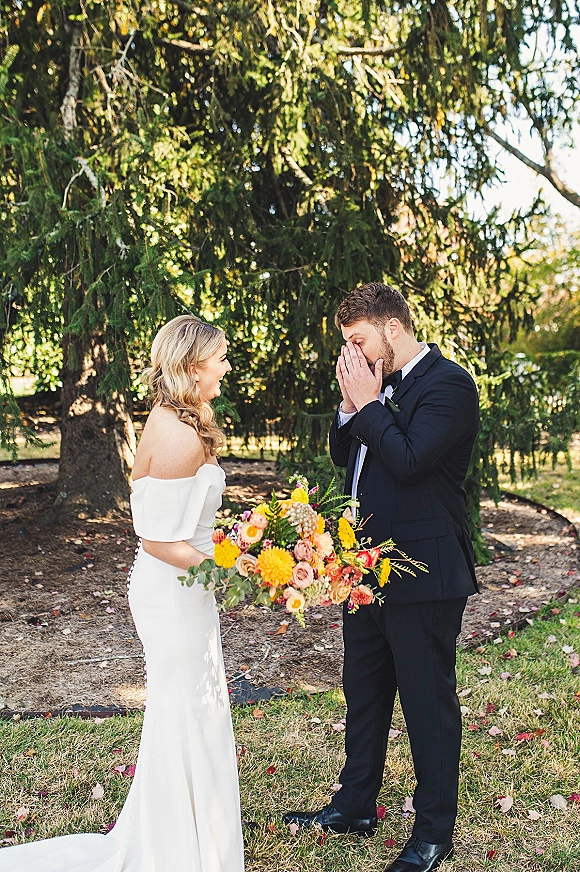 First look moment as bride holds a bouquet and groom in tuxedo reacts on a garden path with evergreen trees and fallen leaves