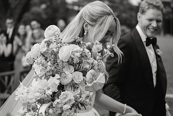 Wedding recessional of bride and groom walking and laughing as guests cheer in a garden, bride holding a bouquet with veil flowing
