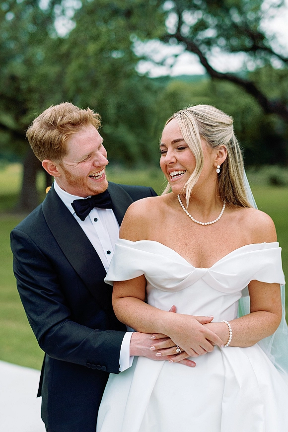 Couple portrait of bride and groom laughing as the groom hugs her, bride in veil and pearls on a green lawn with trees