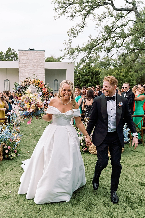 Wedding recessional as bride and groom walking hand in hand past aisle flowers, guests cheering on an outdoor lawn beneath a floral arch