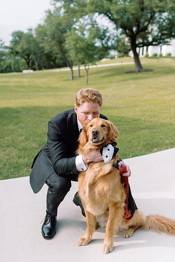 Groom with dog kneeling in a black tuxedo, holding a tuxedo-collared golden retriever on a leash on a grassy lawn with trees