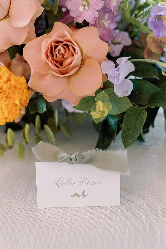 Wedding place card with calligraphy place cards lettering tied in silk ribbon beside a peach rose and purple florals on a linen tablecloth