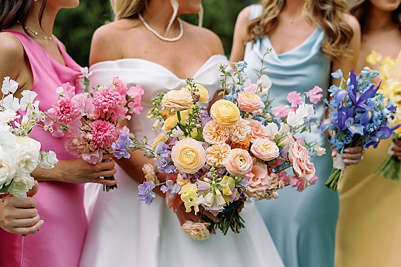 Bridesmaid bouquets in pastel wedding bouquets palette with roses, ranunculus, iris, sweet peas and greenery, held against outdoor greenery background