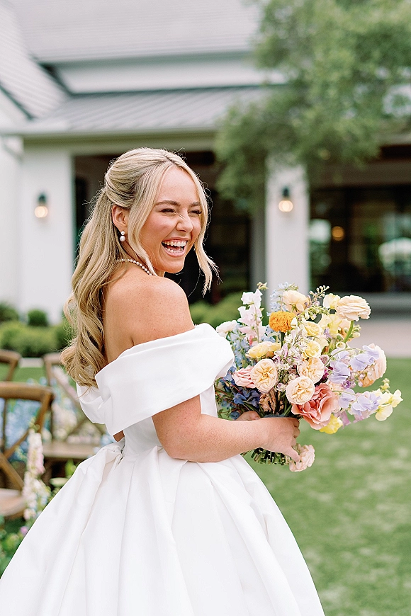 Bridal portrait of a bride holding bouquet, laughing in an off the shoulder gown with pearl necklace on a garden lawn near a white building