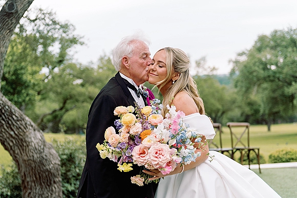 Father daughter portrait as dad kisses the bride’s cheek, her off the shoulder wedding dress and bouquet glowing on a garden lawn with chairs