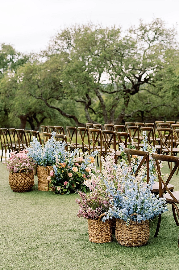 Ceremony aisle decor with meadow flowers in woven baskets beside wood crossback chairs on a grassy lawn under oak trees