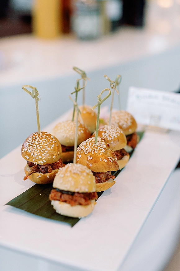 Wedding cocktail bites arranged as mini slider buns with bamboo cocktail picks on a platter lined with banana leaf on a white tablecloth