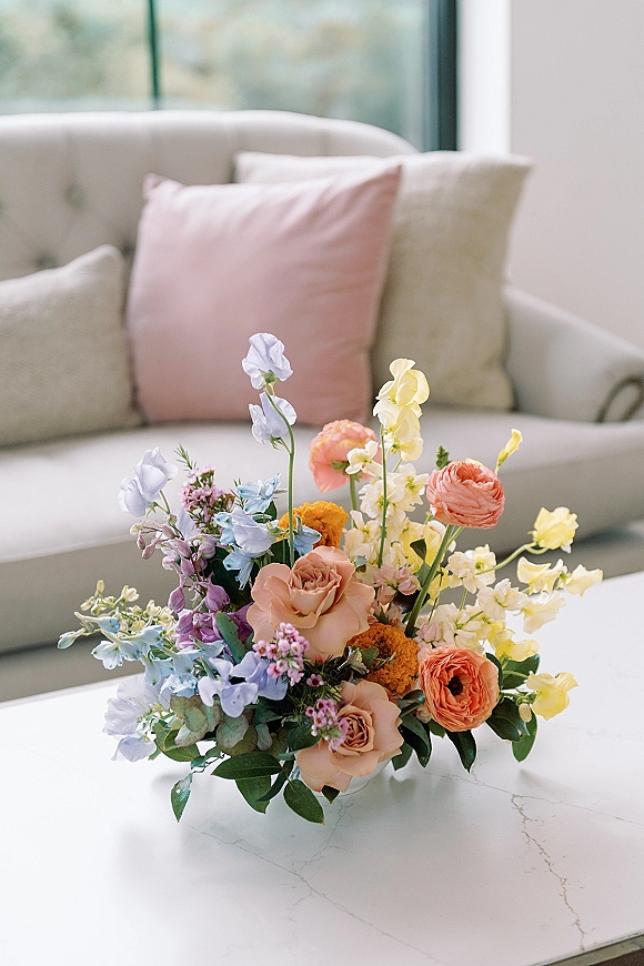Wedding centerpiece of pastel roses, ranunculus, and sweet peas with greenery on a marble tabletop by a window and neutral sofa
