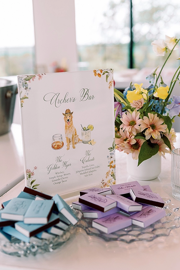 Wedding bar sign with signature cocktail sign illustrations and dog sketch, framed by florals beside pastel blooms on a white tabletop