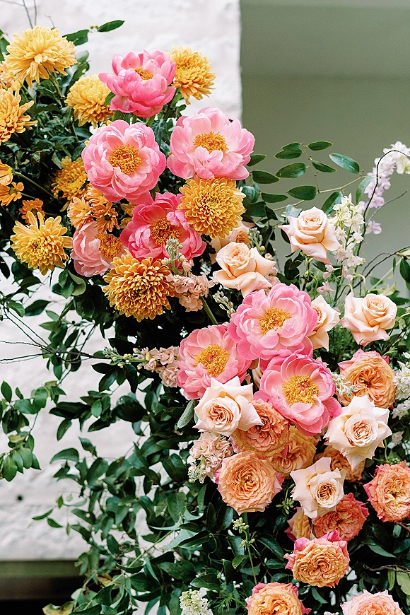 Wedding floral arrangement with peonies and roses, lush greenery, and chrysanthemums set against white and green walls in a bright display