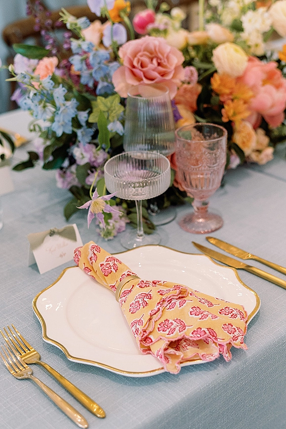 Reception tablescape with wedding table setting featuring pastel floral centerpiece, scalloped plates, gold flatware, coupe glass and place card on linen tablecloth