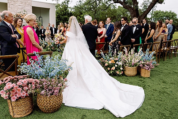 Wedding processional with bride walking down aisle from behind, long veil and train, pastels in woven baskets, guests on lawn under a large tree