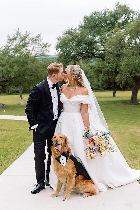 Wedding kiss portrait of bride and groom kissing as their golden retriever in a tuxedo bandana sits beside them on a tree-lined lawn path