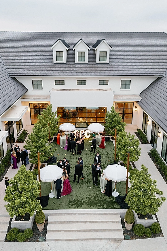 Cocktail hour in a courtyard with outdoor wedding cocktail hour tables under white umbrellas, greenery, and string lights by a white building