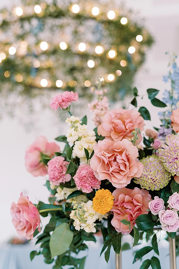 Wedding floral arrangement with a peach rose centerpiece, featuring pink carnations and greenery against a white backdrop with string lights