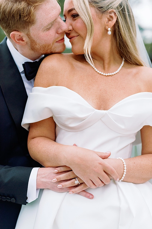 Couple portrait of bride and groom embrace with a gentle forehead touch, her veil and pearl necklace visible against blurred greenery