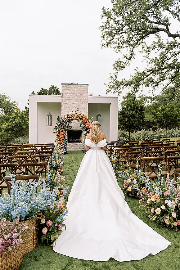 Ceremony setup with an outdoor ceremony aisle leading to a floral ceremony arch by a fireplace, lanterns, and wooden crossback chairs on a lawn