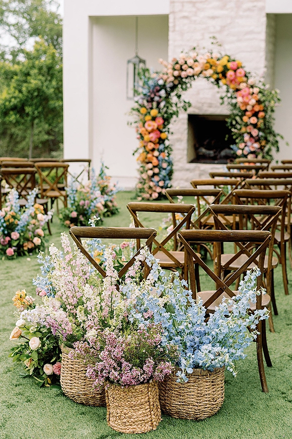 Ceremony setup with an outdoor ceremony aisle of wooden crossback chairs leading to a floral arch on a lawn by a white stone fireplace