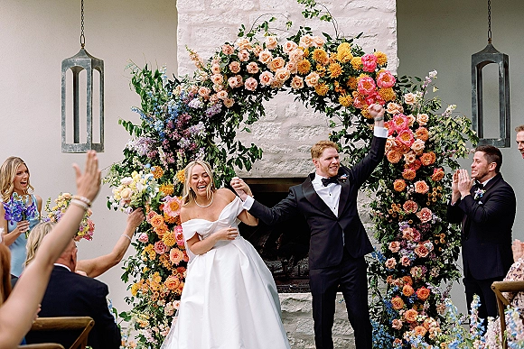 Recessional moment as bride and groom celebrating, holding hands and smiling beneath a rose floral arch with lantern accent indoors by stone fireplace