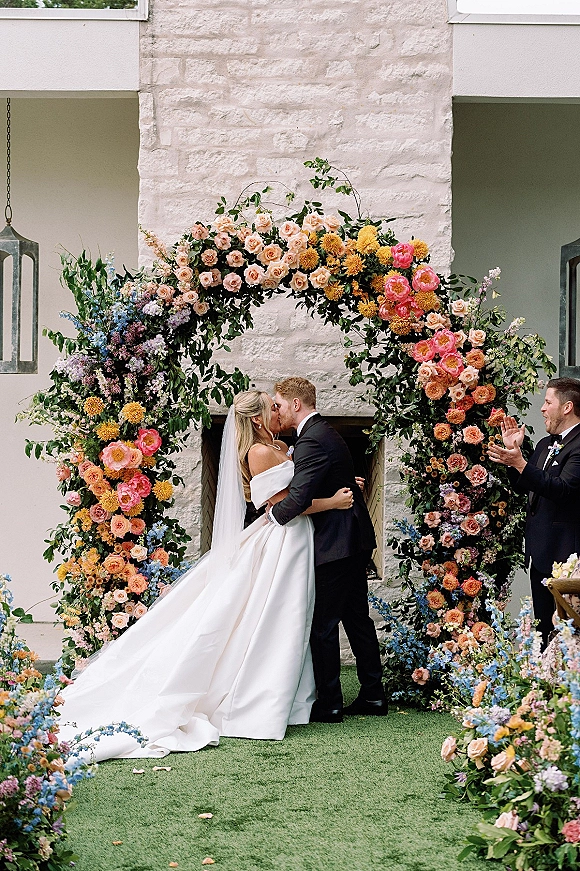 Wedding kiss under a floral arch as the bride in an off the shoulder dress and veil kisses her tuxedoed groom in a courtyard by a stone fireplace