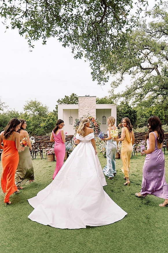 Bride with bridesmaids in colorful bridesmaid dresses walk past aisle flowers and lanterns toward a floral arch on an outdoor lawn