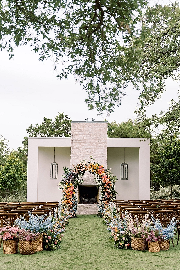 Ceremony setup with an asymmetrical floral arch, aisle flowers in wicker baskets, wooden chairs, and lanterns before a stone fireplace outdoors