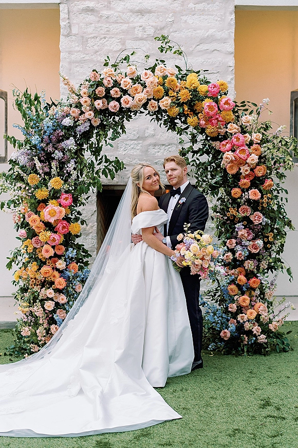 Couple portrait of bride and groom embrace under a round floral arch, her off-the-shoulder gown and veil by a stone fireplace