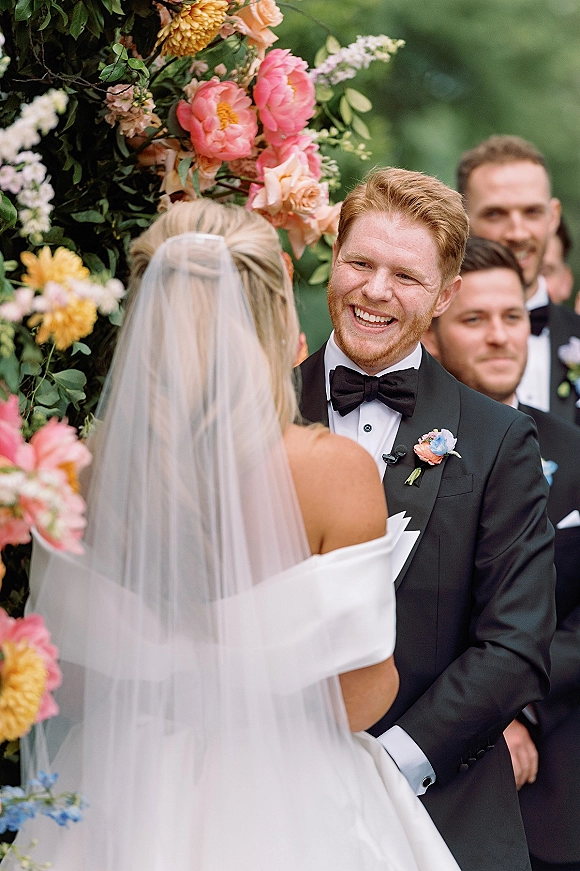 Ceremony moment as bride and groom at altar, groom smiling at bride under a colorful floral arch with groomsmen in greenery behind